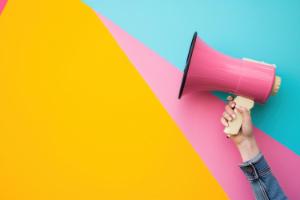 Photograph of arm in denim holding a pink megaphone against a colourful background