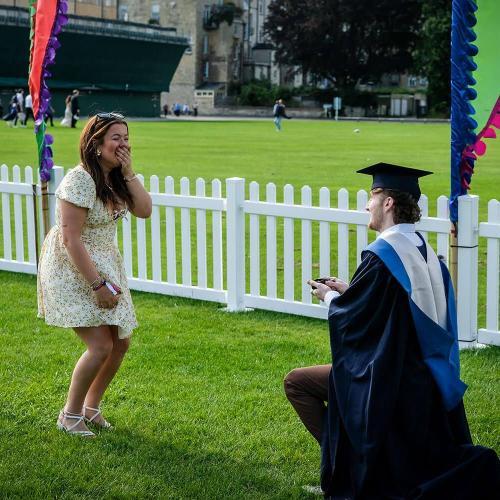 A graduate in ceremonial cap and gown, bends down on one knee in front of a woman in a flowery dress who has her hand over her mouth in surprise. They are in a grass area with festival banners and a picket fence behind them.