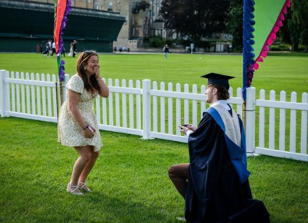 A graduate in ceremonial cap and gown, bends down on one knee in front of a woman in a flowery dress who has her hand over her mouth in surprise. They are in a grass area with festival banners and a picket fence behind them.