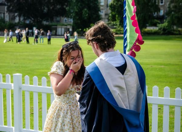 A graduate in ceremonial gown looks towards a woman in a flowery dress who has her hand over her mouth in surprise. They are in a grass area with festival banners and a picket fence behind them.