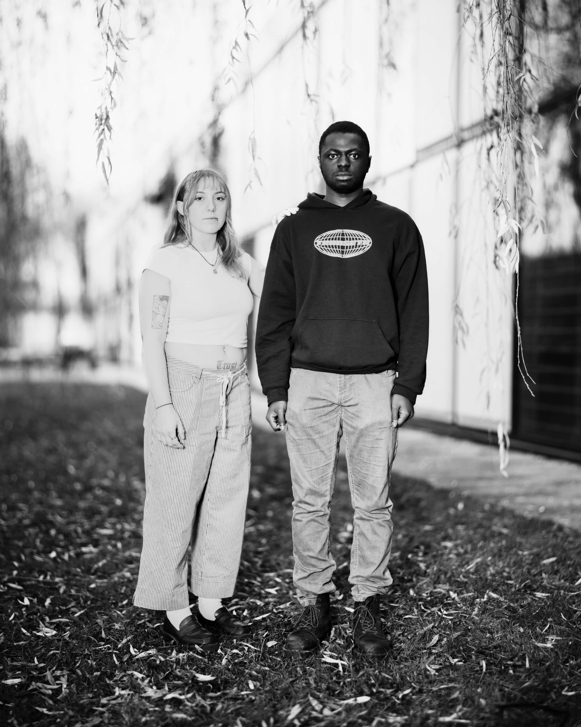 A black and white portrait image of two students stood on a patch of grass outside of a building