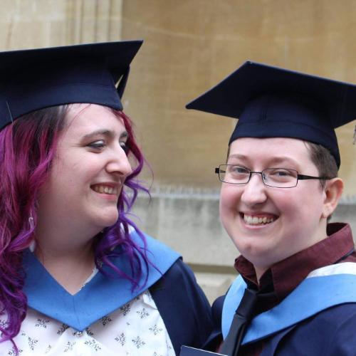 Two graduates stand together in hats and gowns at graduation