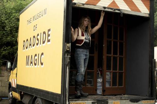 A young woman in jeans and a waistcoat poses from inside the back of a truck
