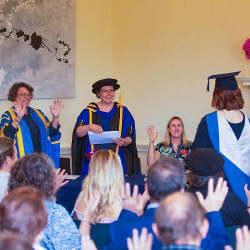 an audience watches and waves hands in silent celebration of a graduate in cap and gown walking up to the front of a room to meet two people in academic regalia and another seated.