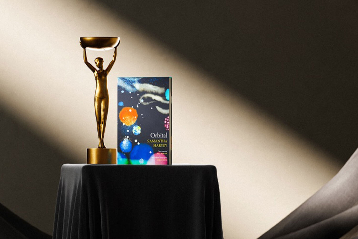 An Image of a book, Orbital, written by Samantha Harvey, on a plinth next to the Booker Prize trophy