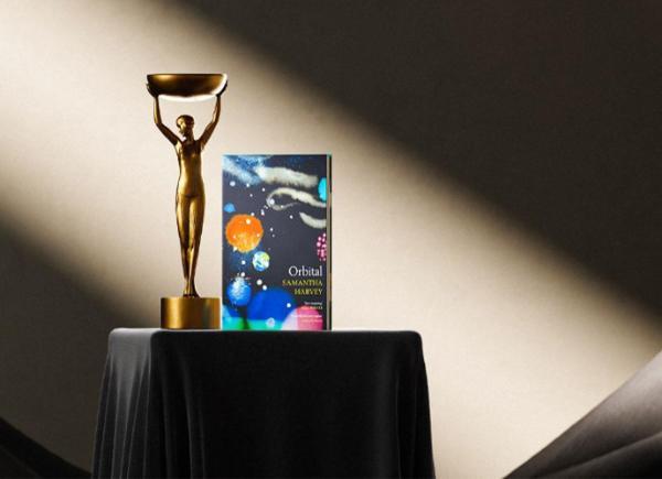 An Image of a book, Orbital, written by Samantha Harvey, on a plinth next to the Booker Prize trophy