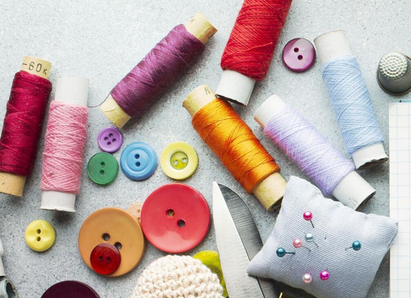 spools of thread, tape measure, scissors, a note pad, buttons and a pin cushion spread out on a white surface.