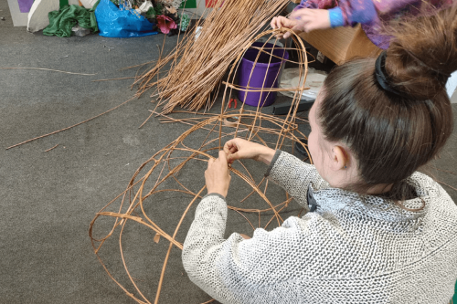 An artist sits on the floor making a sea slug structure out of wicker twigs