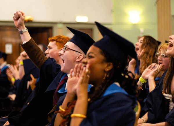 Two rows of students at their final year graduation looking at the stage and cheering.