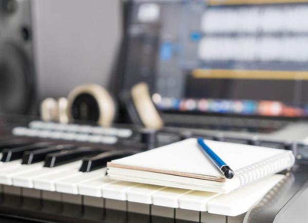 A notebook and pencil rest on top of a keyboard. Monitors showing music editing software, and headphones are blurred in the background.