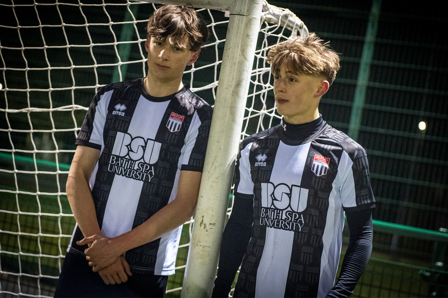 Two young footballers in front of a goal net wearing a bath city + bath spa university shirt