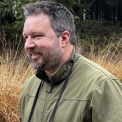 Person with short dark hair and beard in green jacket in field