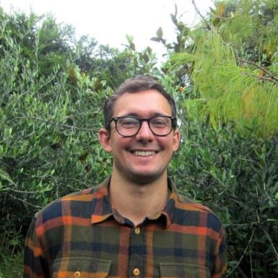 Person with short dark hair and glasses in checked shirt standing in front of high green foliage