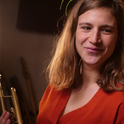 Person with long dark blonde hair in red dress holding a trumpet