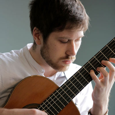 Person with short dark hair and moustache in white shirt playing guitar