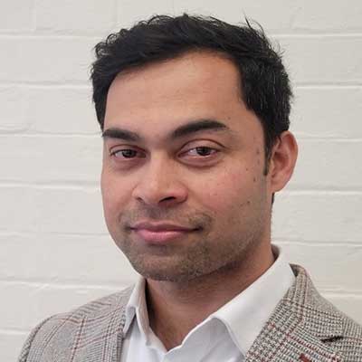 A person wearing a blazer and white shirt stands in front of a white wall