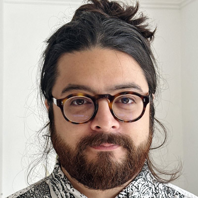 Person with dark hair in a top knot, dark beard and glasses in black and white patterned shirt