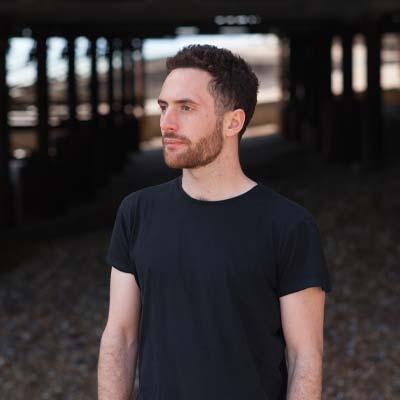 Person with short dark hair and beard in black t shirt on a shale beach under a pier