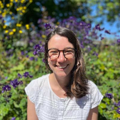 Person with long brown hair wearing glasses and a white sleeveless top smiling in front of purple-flowered shrubs