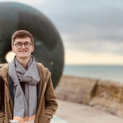 Someone in a beige jacket with big scarf standing on sea shore