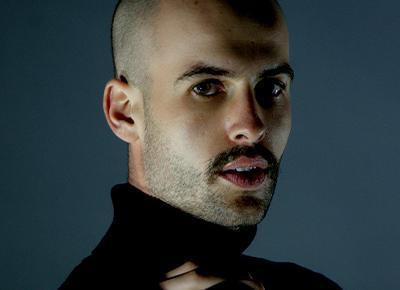 A moody image of an individual with a moustache looking at the camera. 