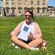 A person sits cross-legged on the lawn in front of Main House, holding their dissertation