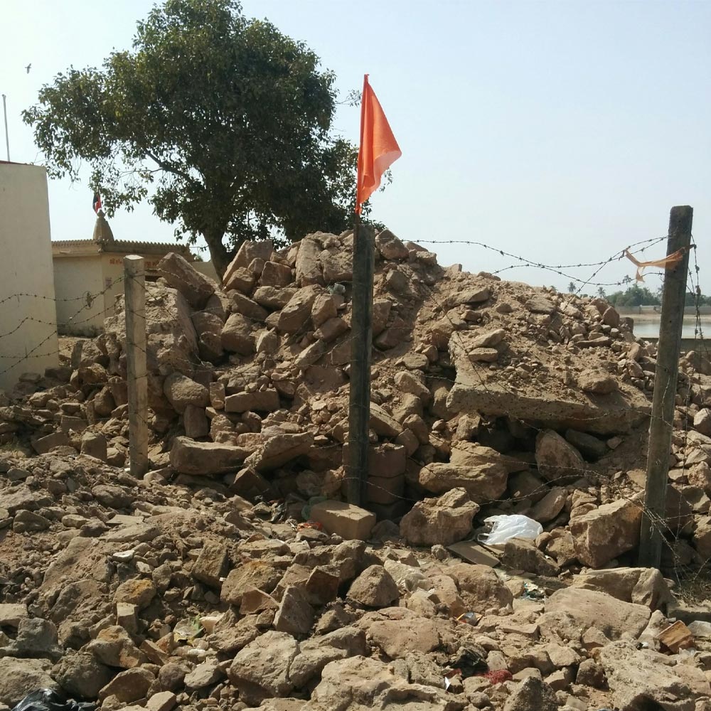 Image of greybrown rubble from a building with a fence and tattered flag. Tree in background.