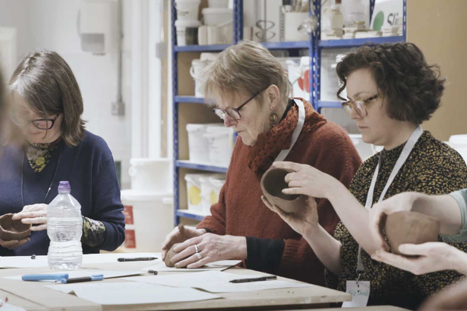 Several people sit around a table and adjust their pottery pieces