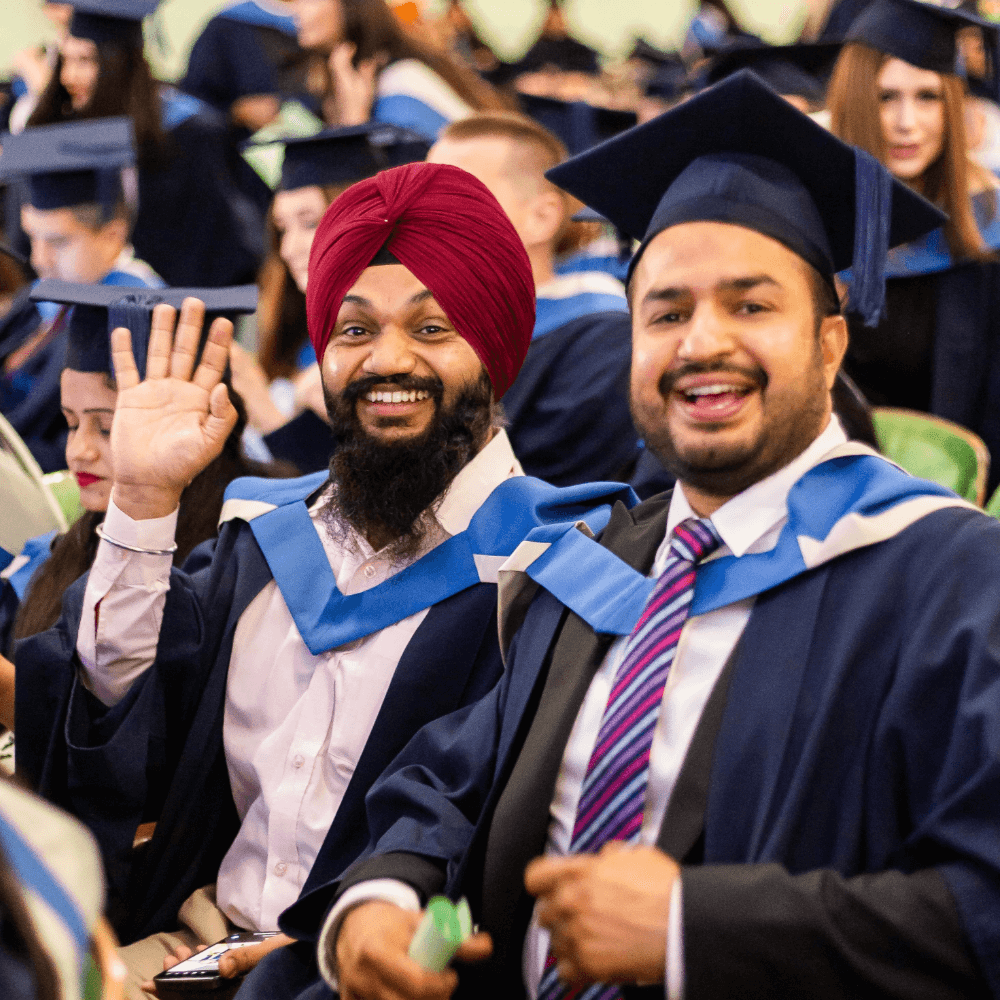 Two students smile and wave to the camera whilst wearing their graduation robes.