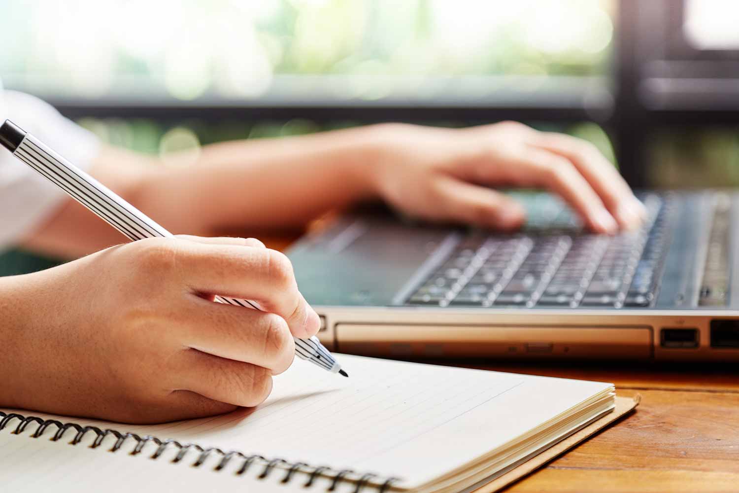 Hands writing in a notepad and typing on a laptop