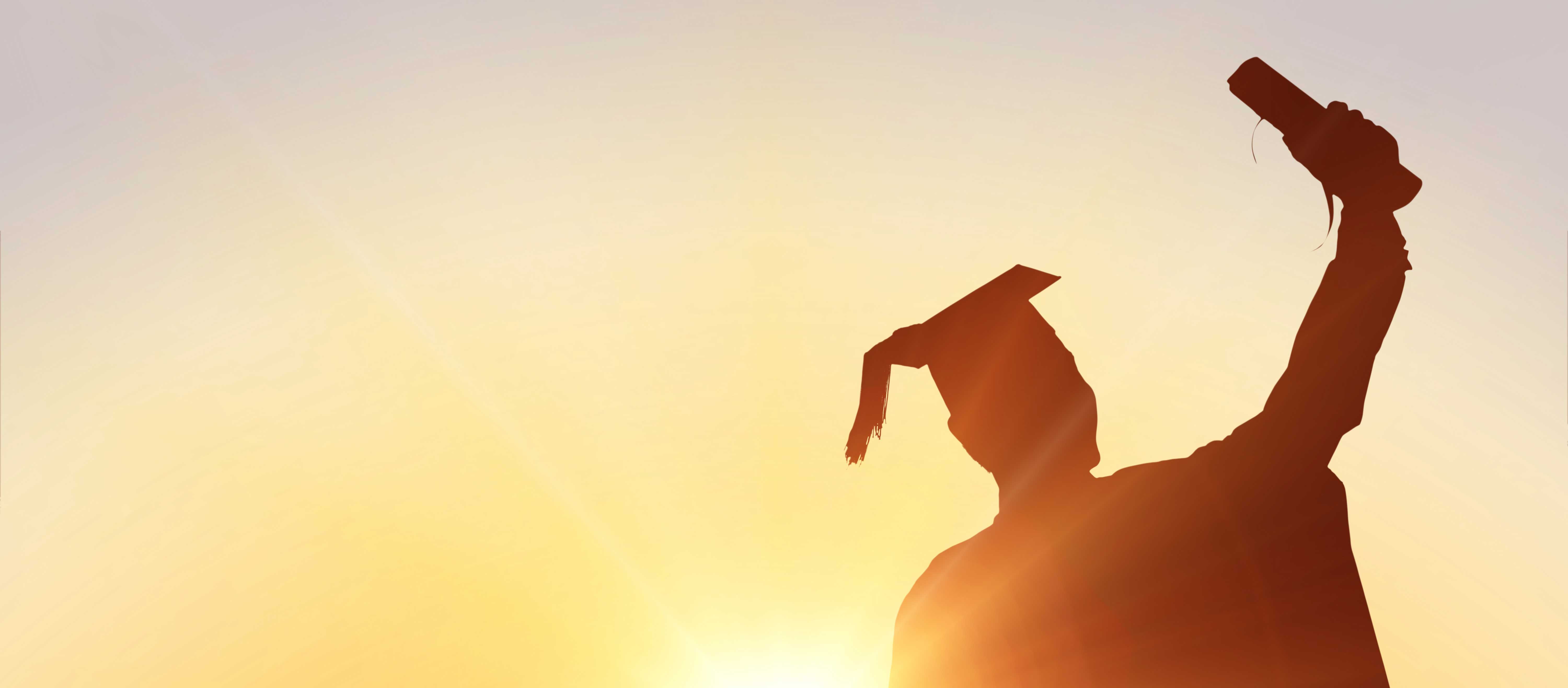 A graduate standing in front of a sunset holds their diploma in the air