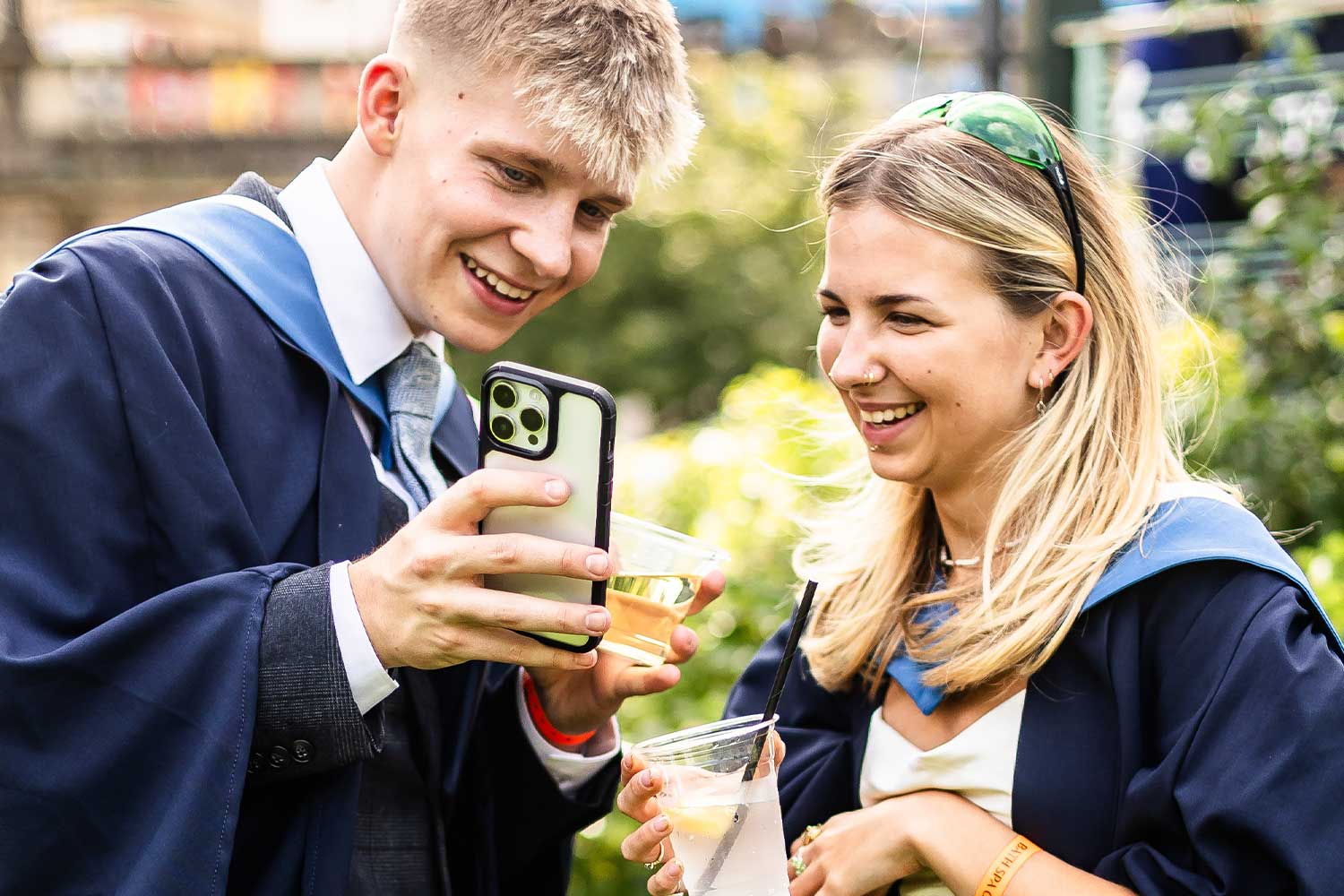 Two graduates holding drinks in plastic cups look at an iPhone