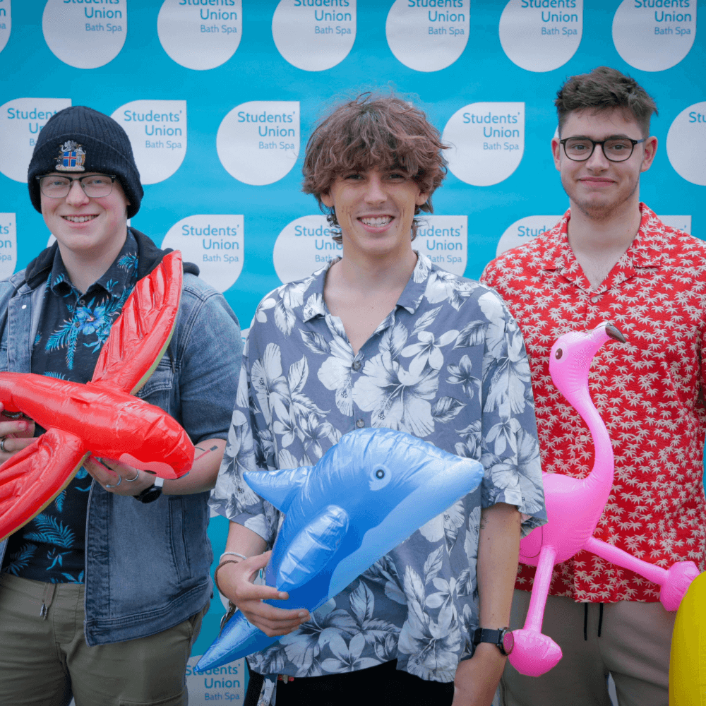 Three students with inflatable toys stand in front of a Students Union branded backdrop, smiling.