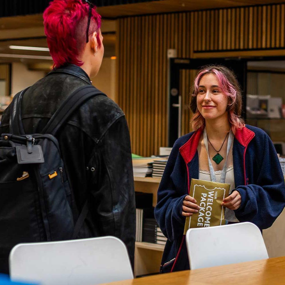 Two students having a conversation in university setting. One holding a Welcome to Uni folder