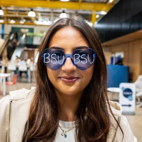 A person with long brown hair wears blue heart-shaped sunglasses that say 'BSU' on each lens