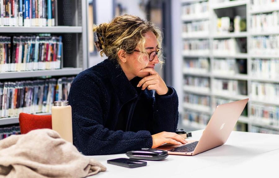 Student working on a rose gold laptop in a library, with shelves of books behind her and a phone, water bottle and cardigan nearby on the table